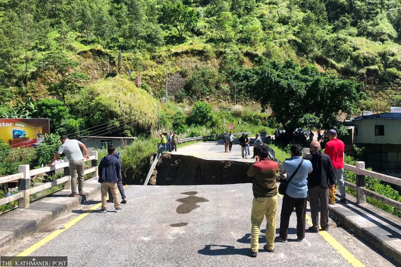A damaged road with a collapsed section, surrounded by people observing the situation.